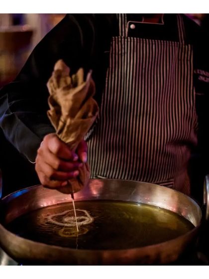 A chef pipes concentric circles of batter into hot oil to create our signature nano crispy jalebis. This action shot highlights the skill and traditional technique involved in making this beloved sweet.