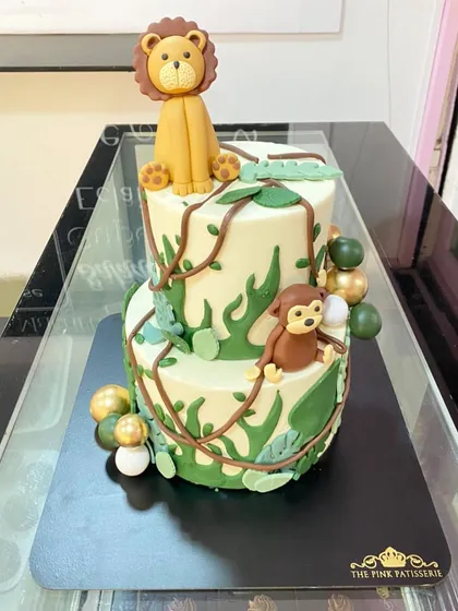 A two-tiered jungle cake with a lion on top and a monkey climbing the side. The cake is decorated with fondant vines and leaves to create a lush jungle feel.