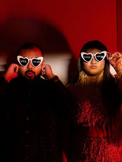 A fun and quirky portrait of the couple posing with heart-shaped sunglasses under dramatic red lighting, showing their playful side during the after-party celebrations.