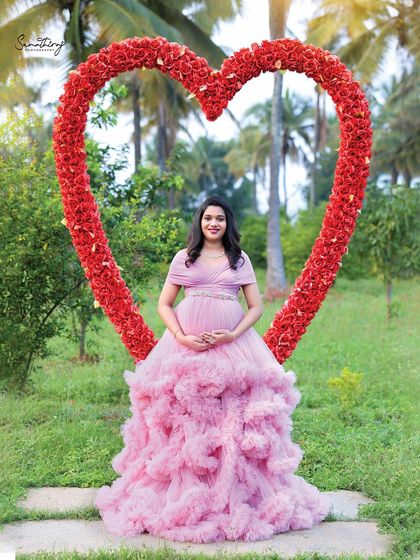 A solo portrait of a mother-to-be framed by a large heart-shaped floral prop. The pink ruffled gown and the romantic outdoor setting create a perfect fairytale moment.