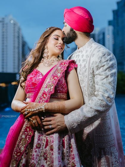 An intimate portrait of a Sikh couple in Bangkok, the groom kissing his bride's forehead with the city's modern skyline and a pool in the background.