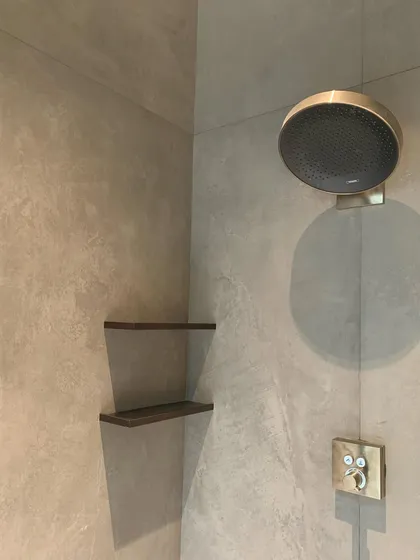 A detail of a shower, showing the contrast between a micro-concrete wall finish and a brushed gold fixture. This juxtaposition of raw and refined materials creates a rich, sensory experience.