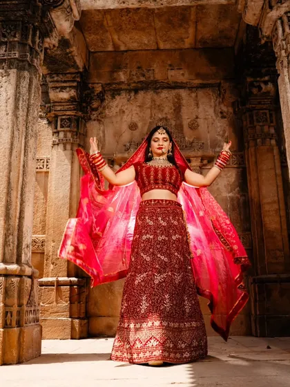 A stunning portrait of the bride in her vibrant red lehenga. With her dupatta flowing, she stands like a queen amidst the ancient stone pillars of the haveli.