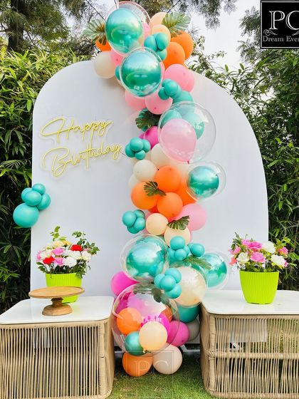 A close-up of the modern and cheerful birthday backdrop. The white arch panel with a neon 'Happy Birthday' sign is adorned with a vibrant balloon garland in pink, orange, and teal.