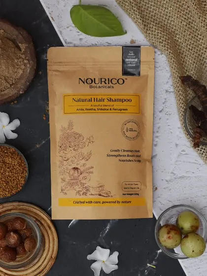 This flatlay tells the story of an Ayurvedic hair care brand through its raw ingredients. Amla, Reetha, and Shikakai are arranged around the product on a textured surface, emphasizing its natural and organic origins.