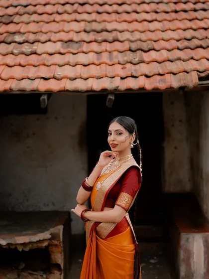A wide shot that places the model in a rustic, traditional setting. The tiled roof and weathered walls of the old house add a story and context to her beautiful saree.