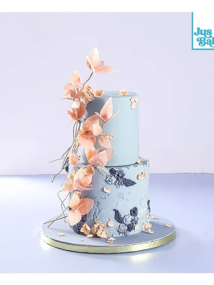 This two-tier designer cake showcases a unique combination of textures and colors. The gray concrete-style bottom tier contrasts beautifully with the smooth blue top tier, all brought together by delicate, handmade wafer paper flowers.