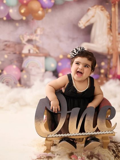 A glamorous first birthday portrait with a carousel theme. The combination of a little black dress, pearls, and a sparkling "One" sign creates a truly special image.