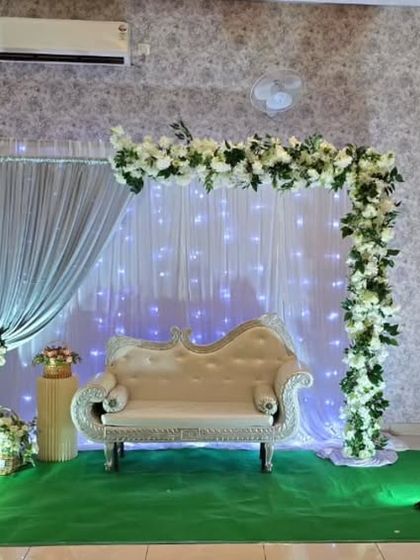 A simple and elegant stage setup with a white floral L-shaped arch, fairy lights, and a classic diwan.