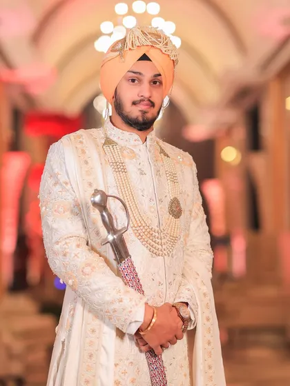 A solo portrait of the groom in his white sherwani and peach-colored turban, holding a kirpan. This image highlights the grace and tradition of a Sikh groom's attire.