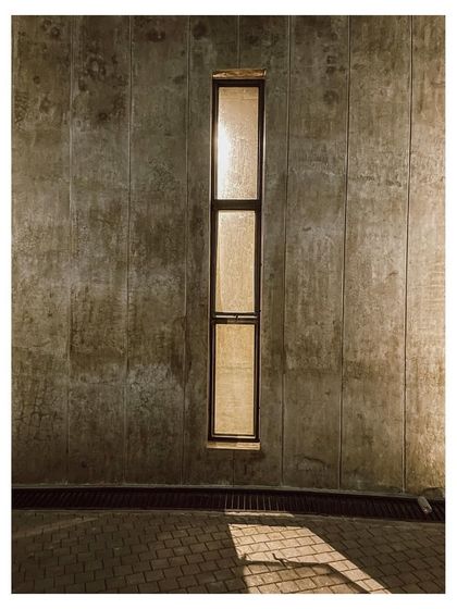 A detail of a narrow window set into the textured concrete wall, creating a sliver of light and a connection to the outside.