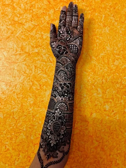 A stunning full-arm bridal mehendi with high-contrast, dense patterns. This design showcases a mix of floral, paisley, and geometric elements, creating a dramatic and unforgettable look against the bright yellow background.