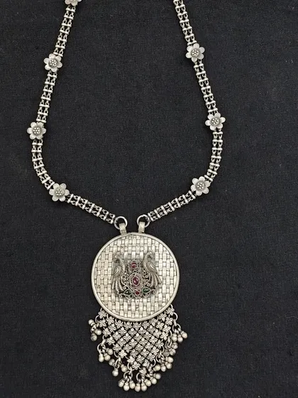 An antique-style oxidised silver necklace. The design features a floral chain and a central pendant with a peacock motif and a dangling net of silver beads.