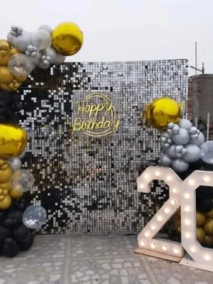 An outdoor 20th birthday party on a terrace. This setup includes a silver and black sequin wall, a black and gold balloon garland, and marquee numbers.