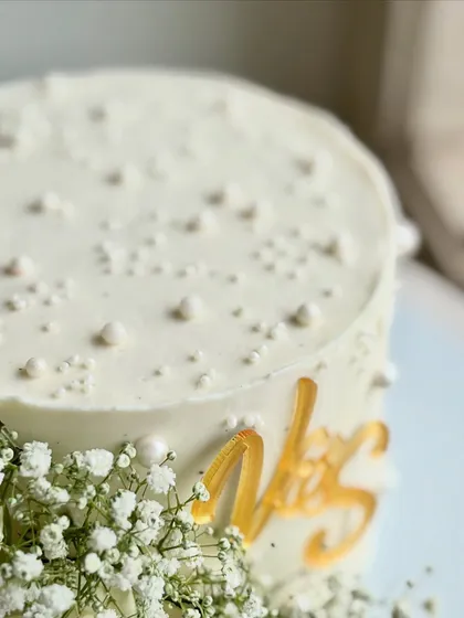 A macro shot focusing on the texture of the buttercream, the tiny edible pearls, and the custom gold initials, capturing the cake's minimal yet luxe vibe.