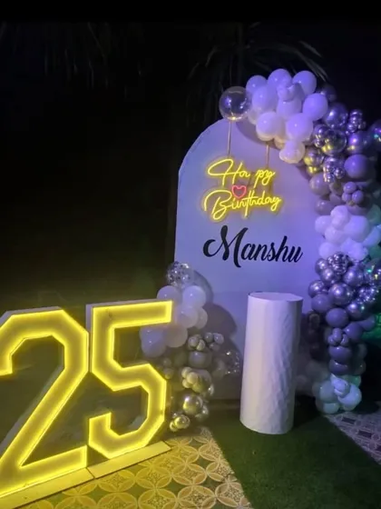 An outdoor 25th birthday party at night. The setup features a purple and silver balloon arch against a custom backdrop, with large illuminated numbers.