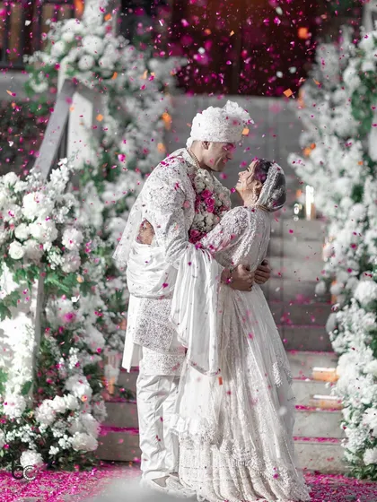 An all-white wedding dream come true. The couple shares a loving embrace as a shower of pink petals rains down around them, framed by elegant white floral decor.