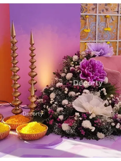 A close-up of the decor for a Mehendi ceremony, showing traditional gold 'diyas' and bowls of marigold petals next to a vibrant floral arrangement.