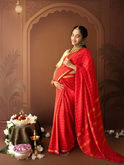 A graceful standing portrait of a mom-to-be in a red saree, holding a peacock feather, with a traditional backdrop adding to the cultural richness.