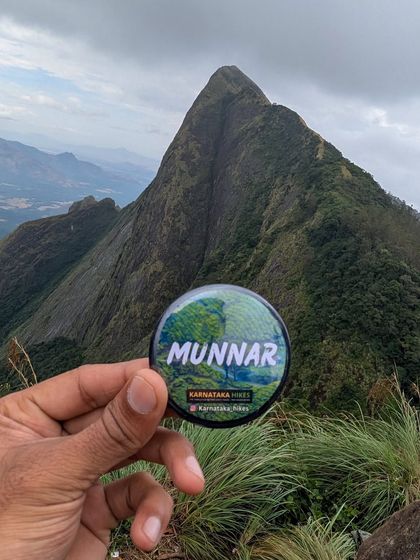 Our Munnar trip badge held up against a stunning, sharp peak, a perfect souvenir of the adventure.