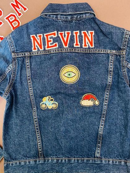 A custom denim jacket for 'Nevin', featuring his name in bold red letters. The jacket is decorated with unique patches, including an evil eye, a bicycle, and a Santa hat.