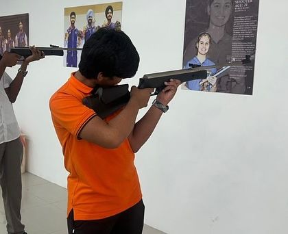 Students practice their aim with air rifles against a wall featuring posters of shooting icons. We aim to inspire the next generation by connecting them with the heroes of the sport.