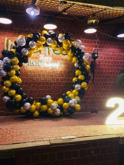 A cool and edgy 25th birthday decoration against a rustic brick wall. The full ring of black, gold, and silver balloons, combined with a neon sign and large marquee numbers, creates a high-impact look for a memorable party.