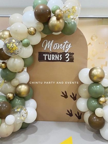 A subtle and stylish Jurassic theme for a third birthday. The decoration uses earthy tones of green, brown, and beige balloons with dinosaur footprint details on the backdrop.