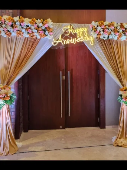 An elegant entrance arch designed for a 25th wedding anniversary. It features soft golden drapes, beautiful floral arrangements, and a glowing neon 'Happy Anniversary' sign to welcome guests.