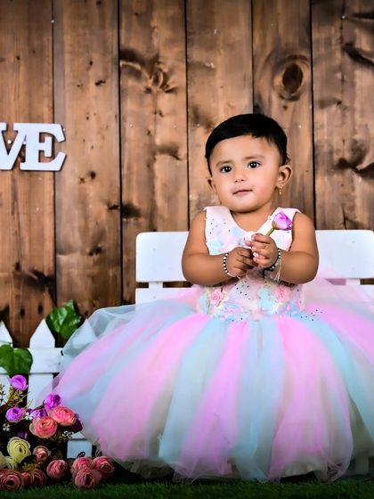 A baby girl in a beautiful pastel dress sitting by a white picket fence.