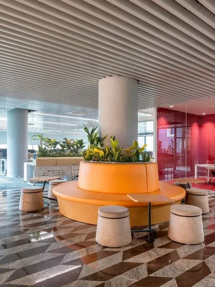 The central collaborative hub at the Pinnacle campus features a vibrant, biophilic design with an orange circular seating area and integrated planters. This space is designed to foster spontaneous interaction and community among employees, with geometric flooring and modern finishes.