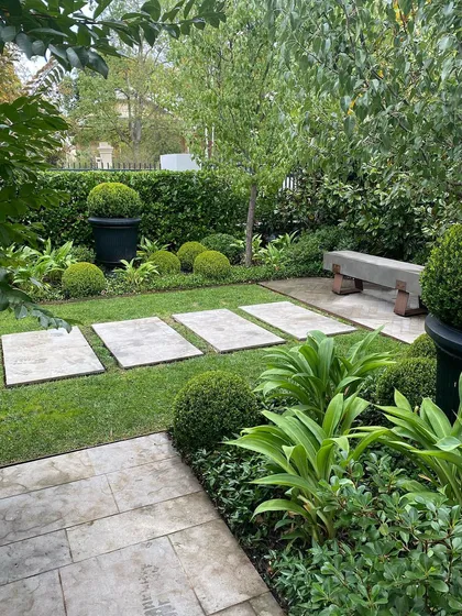 A serene corner of a garden featuring a stone bench and a path of stepping stones set in the lawn. The space is filled with a mix of formal Buxus spheres and lush, leafy plants.
