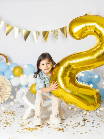 A series of portraits celebrating a second birthday. The toddler poses with a giant gold '2' balloon, surrounded by blue and white balloons and confetti for a festive look.
