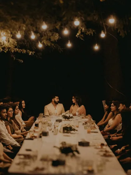 An atmospheric shot of the couple at their engagement dinner, seated at the head of a long table under hanging lights.