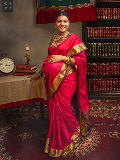 A portrait that evokes a sense of history and intellect. The mother-to-be, in a rich red saree, stands in a library of old books, symbolizing the wisdom and stories she will pass on.