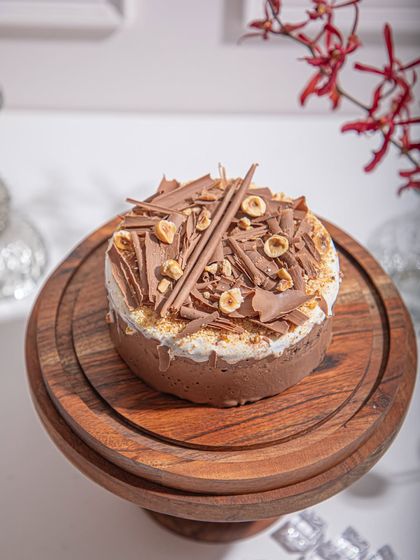 Our Black Forest Ice Cream Cake on a wooden stand, topped with chocolate shavings and hazelnuts. A perfect dessert for birthdays or just because.