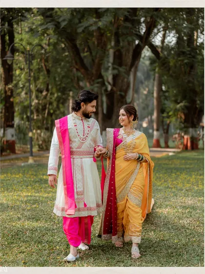 A candid walking shot from a Marathi-themed pre-wedding session. The couple's coordinated outfits, with the groom's white sherwani and pink stole matching the bride's saree, create a visually cohesive and beautiful image.