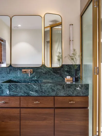 The master bathroom combines rich materials for a luxurious feel. A vanity crafted from green Indian marble is paired with dark wood cabinetry and elegant brass-framed mirrors, creating a sophisticated and functional space.