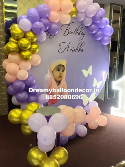 A personalized first birthday celebration with a custom photo backdrop. This butterfly-themed setup in pastel purple and pink is complemented by a large light-up marquee number '1' and golden balloons.