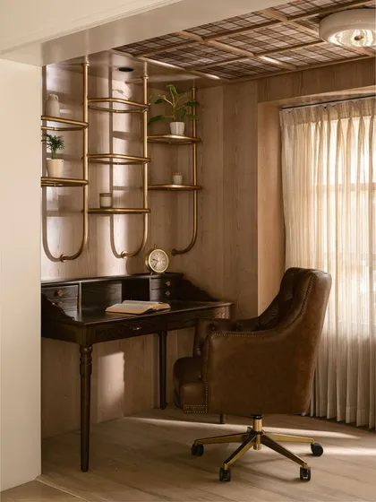 This home office features a classic dark wood secretary desk paired with a tufted brown leather office chair. Above, a custom-designed sculptural brass shelving system adds a modern, artistic touch.
