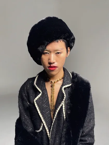A close-up portrait from the shoot, focusing on the model's bold red lip, fur hat, and gold chain necklace. The styling is opulent and confident.