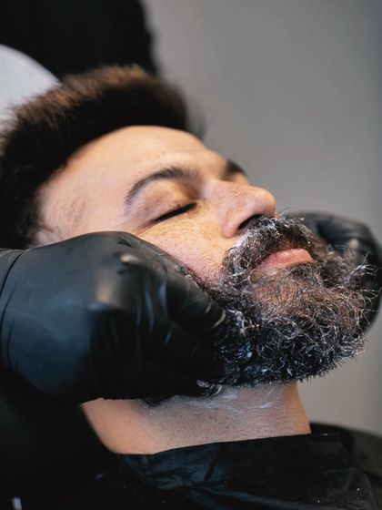 A close-up of a client receiving a beard grooming service. The stylist is applying shaving cream, preparing for a precise shape-up.