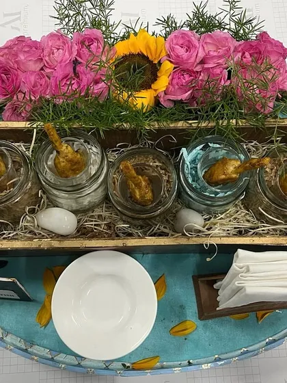 A rustic yet elegant presentation of various dips or small bites in glass jars. The arrangement on a bed of straw within a wooden crate, adorned with pink roses, creates a charming and memorable serving style.