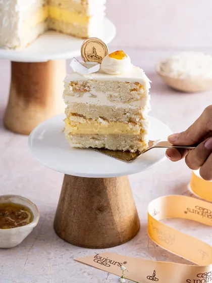 A hand lifting a slice of our Coconut & Passionfruit cake, showing its light and airy texture. The layers of coconut sponge and passion fruit filling are perfectly balanced for a refreshing, tropical dessert experience.
