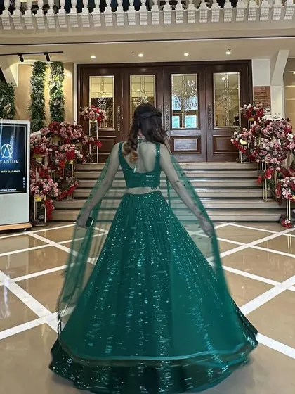 A client twirling in a stunning emerald green sequined lehenga in a grand hotel lobby. The cape-style dupatta adds a modern and dramatic flair.
