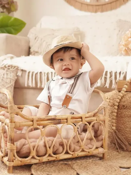 A playful shot of a baby boy tipping his hat while sitting in a wicker bassinet during his charming, boho-themed first birthday shoot.