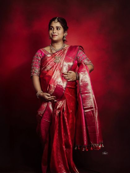A stunning full-length portrait of a mother-to-be in a radiant red silk saree. The rich color and studio lighting beautifully highlight the texture of the fabric and the joy of expectancy.