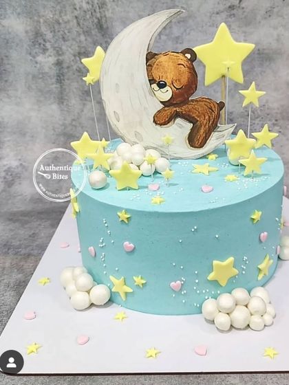 A dreamy baby shower cake with a teddy bear sleeping on the moon. The light blue cake is decorated with yellow stars and fluffy clouds.