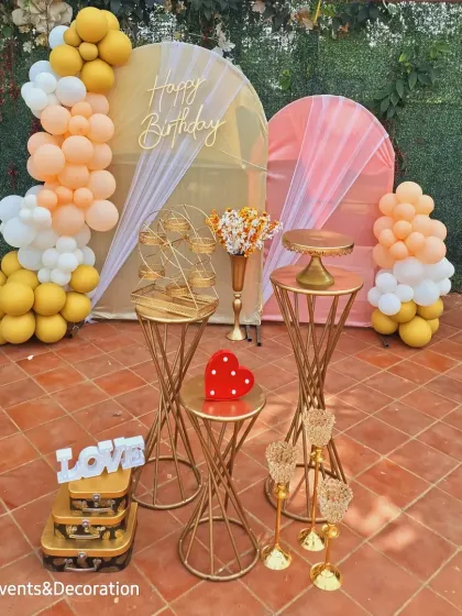 A lovely outdoor birthday setup against a green wall. The dual arched backdrops with soft draping and a pastel balloon garland create a beautiful, airy feel.