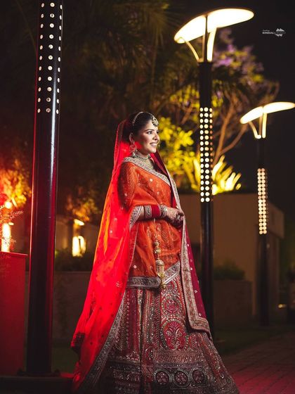 A smiling bride in her vibrant orange lehenga, posing outdoors at night. The modern lighting of the venue contrasts beautifully with her traditional attire.